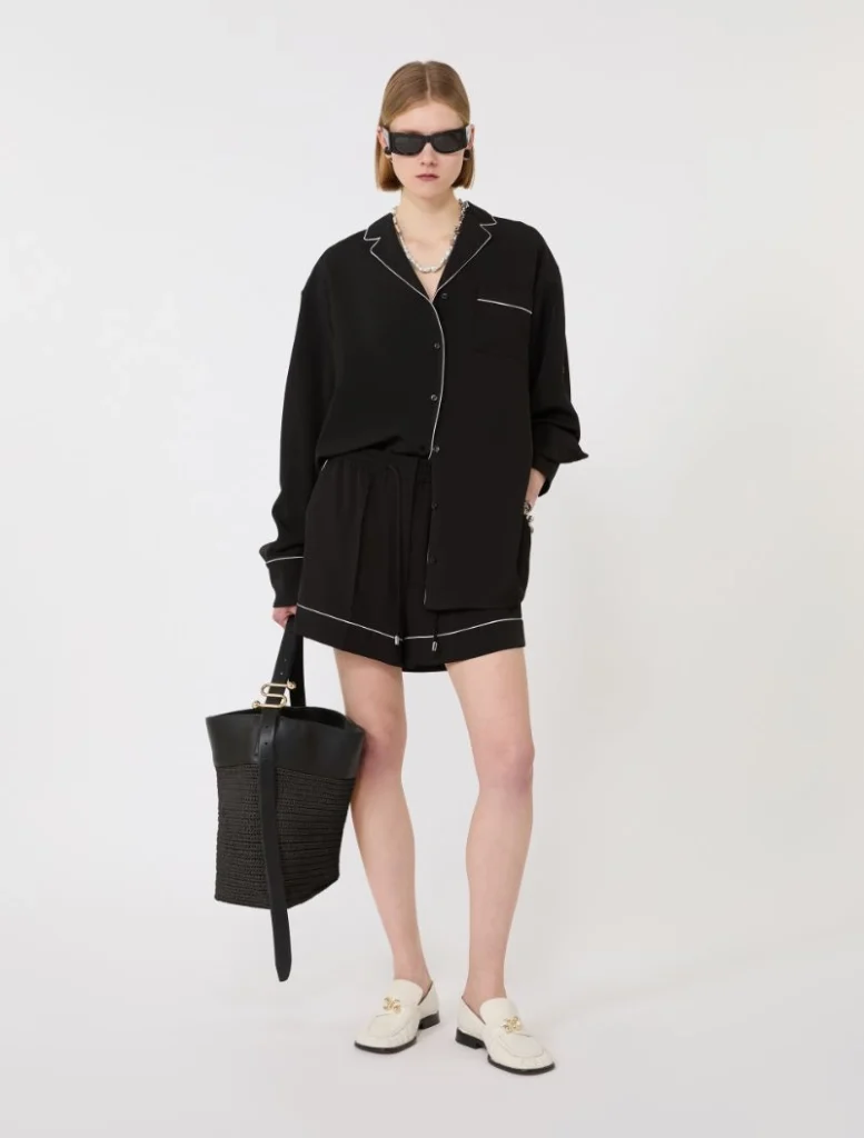 Model wearing a black pajama-style shirt and shorts with white piping, cream loafers, oversized sunglasses, and a black woven tote bag.