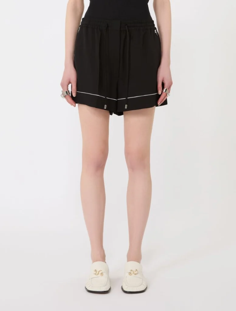 Black athletic-style shorts with an elastic drawstring and white piping, paired with white loafers, shown from the waist downening the legs.