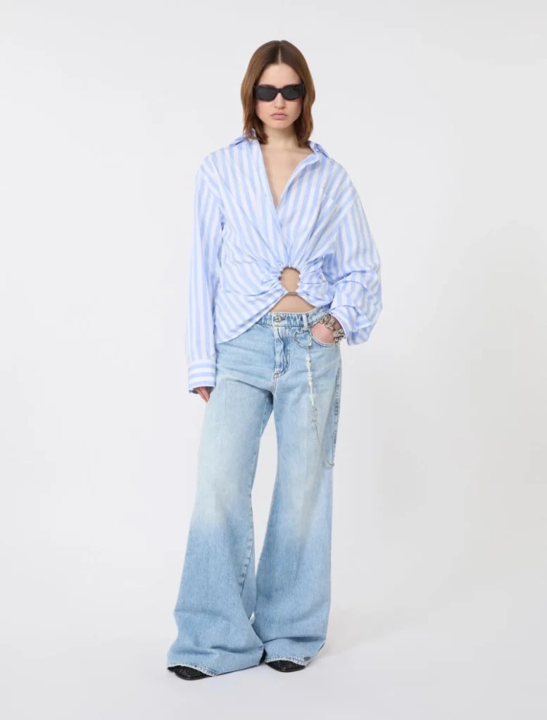 Model wearing a blue-and-white striped shirt tied at the waist, with wide-leg light-wash jeans and black sunglasses.