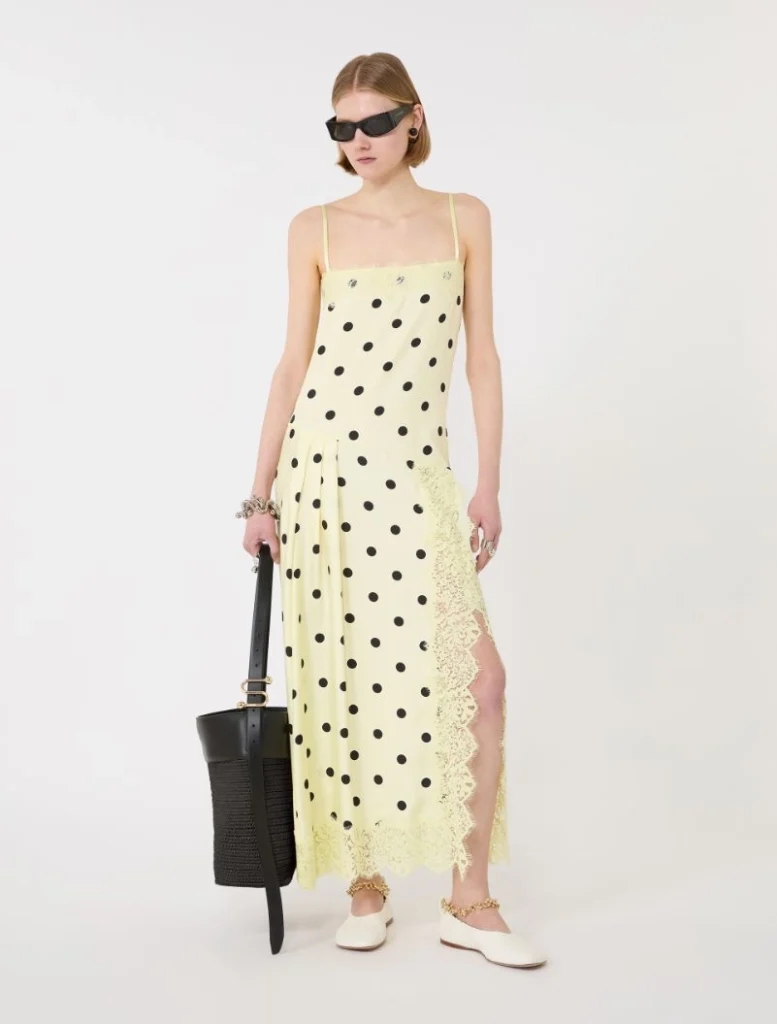 Model wearing a pale yellow spaghetti-strap dress with black polka dots and lace trim, holding a black woven tote and wearing white flats with sunglasses.