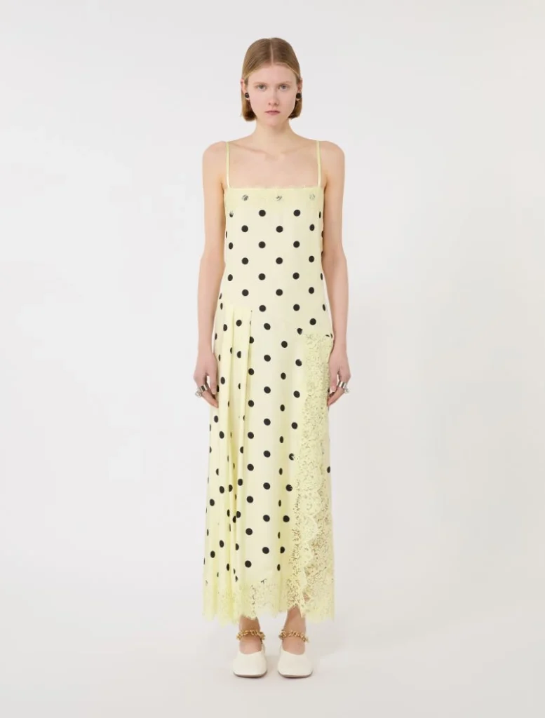 Young woman wearing a pale yellow spaghetti-strap maxi dress with black polka dots and lace trim at the hem, standing against a light background.