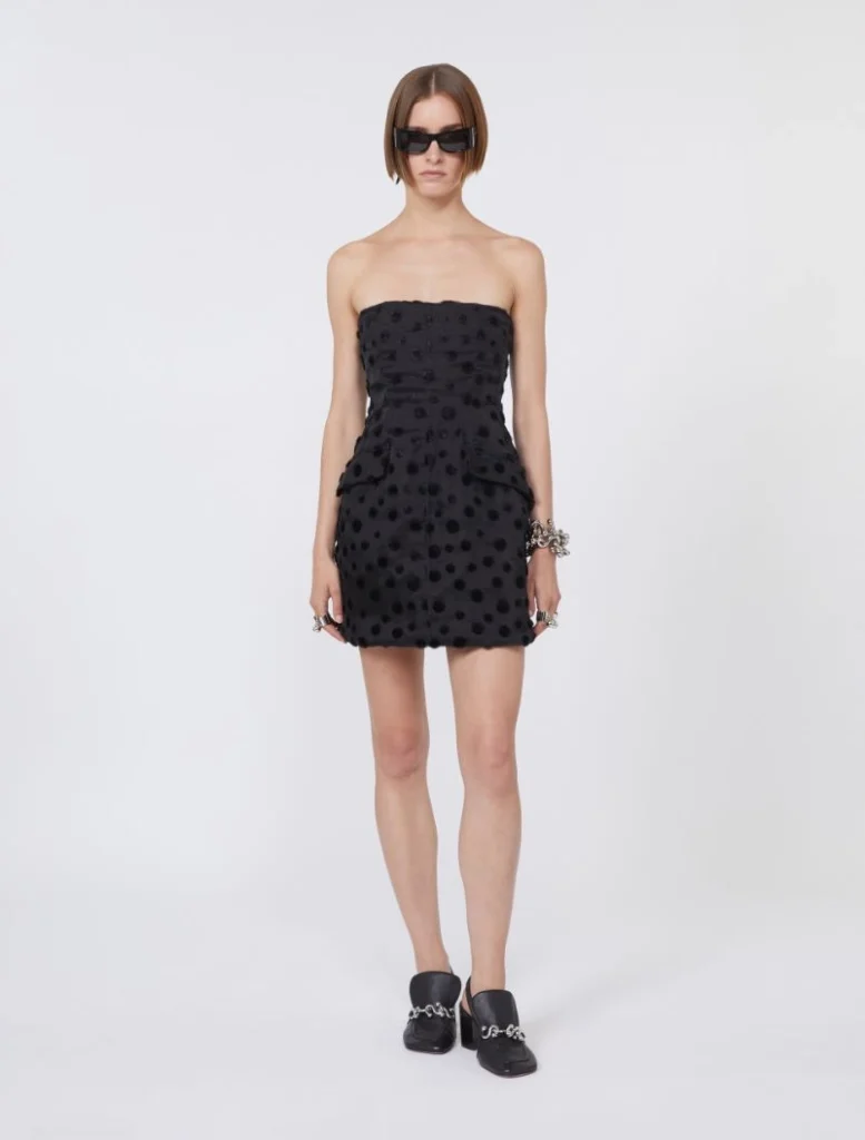 Female model in a strapless black mini dress with polka-dot texture, paired with black sunglasses and chunky bracelets, wearing black chain-detail loafers.