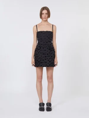 Standing female model wearing a black polka-dot mini dress with thin straps and side pockets, paired with black loafers with chain detail.