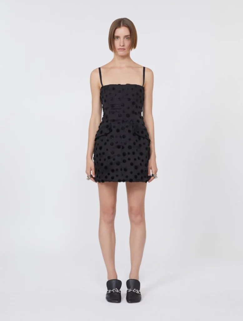 Standing female model wearing a black polka-dot mini dress with thin straps and side pockets, paired with black loafers with chain detail.