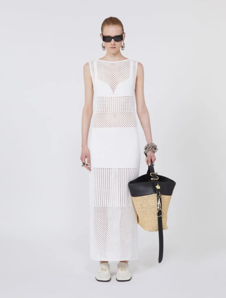 Model wearing a white perforated maxi dress, black sunglasses, and chunky bracelets, holding a straw tote bag.