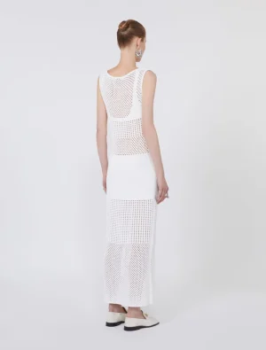 Back view of a woman in a white sleeveless crochet-knit maxi dress with open-weave panels, wearing a bun and statement earrings.