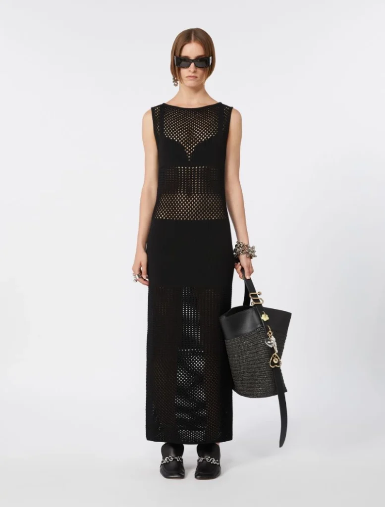 Model in a long black mesh dress with sunglasses, holding a black handbag and wearing chunky jewelry.