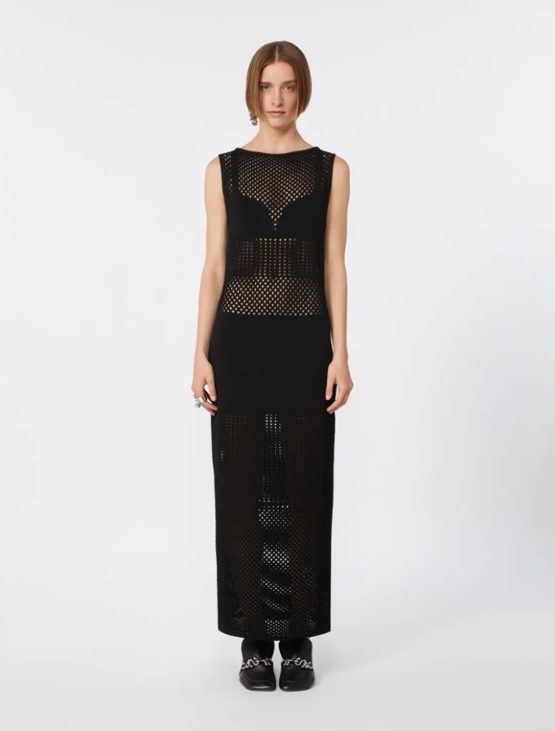 Full-length black mesh maxi dress with perforated panels worn by a female model, paired with black loafers and a simple updo.