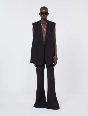 Model in an all-black sleeveless blazer and wide-leg pants, with chunky silver jewelry and dark sunglasses against a white backdrop.