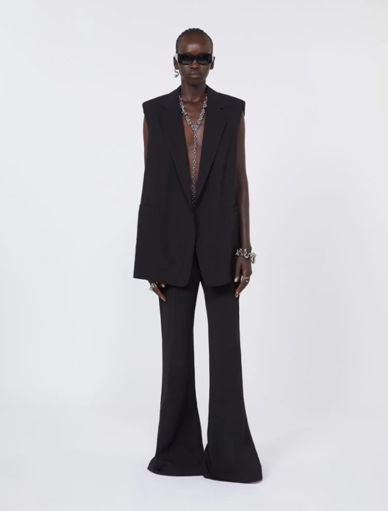 Model in an all-black sleeveless blazer and wide-leg pants, with chunky silver jewelry and dark sunglasses against a white backdrop.