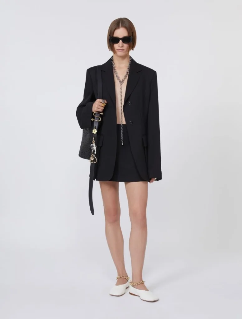 Model in an oversized black blazer with a black mini skirt, white flats, and bold jewelry, wearing dark sunglasses and carrying a black handbag.
