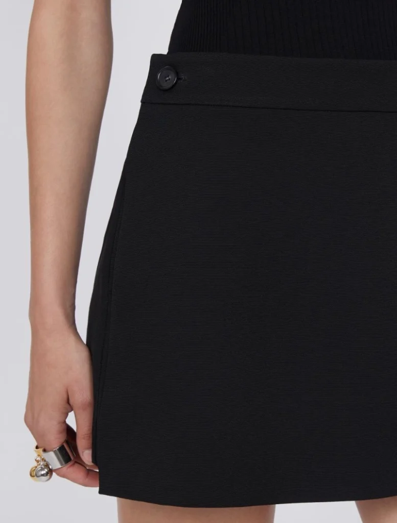 Close-up of a black skirt waistband with a decorative side button; a hand with rings rests at the hem.