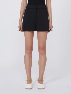 Front view of a person wearing high-waisted black shorts and white flats with gold chain ankle jewelry.