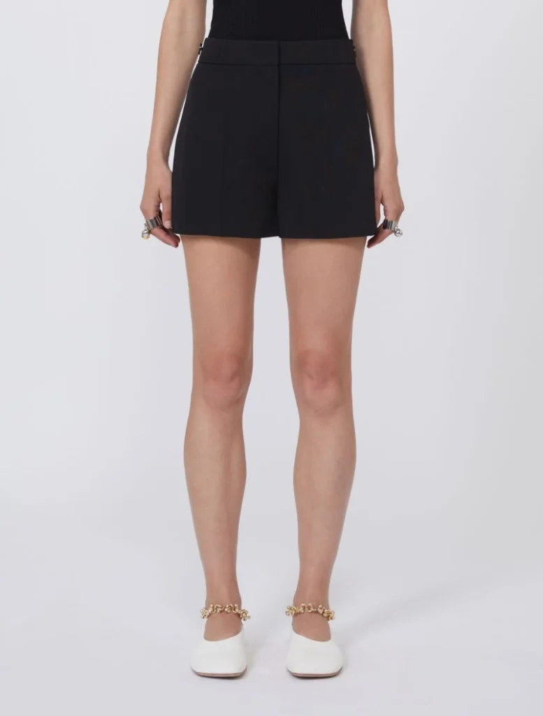 Front view of a person wearing high-waisted black shorts and white flats with gold chain ankle jewelry.