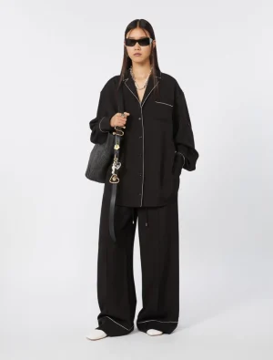 Person in an oversized black pajama-style suit with white piping, black sunglasses, and a gray bag, posing for a fashion shoot.