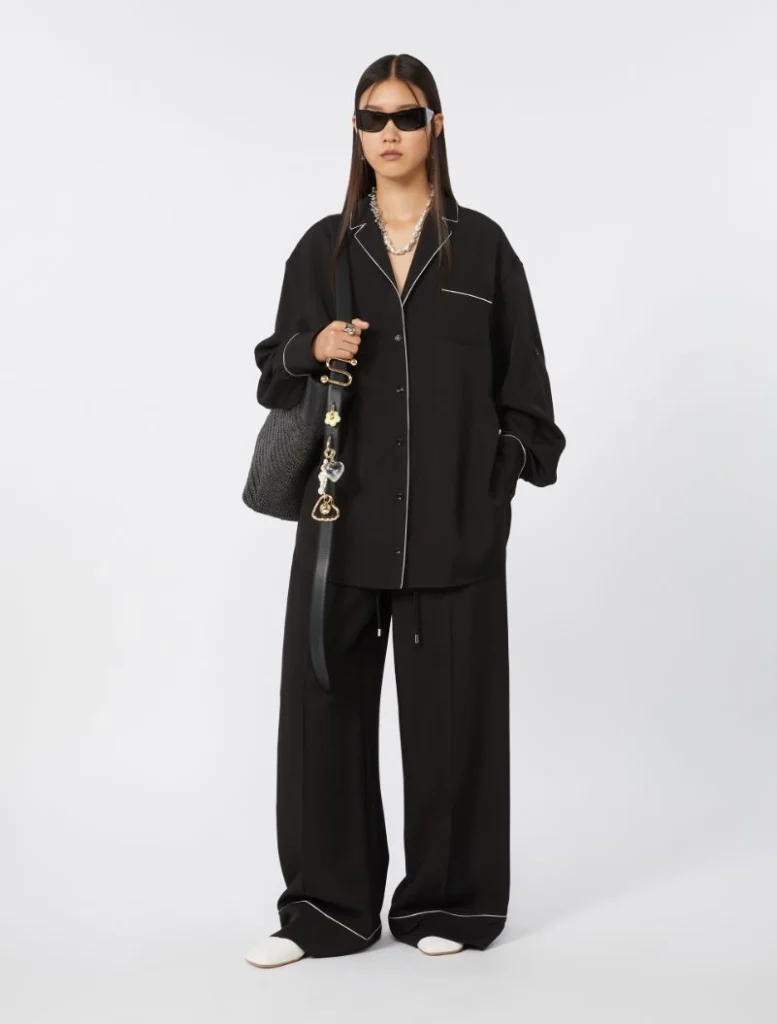 Person in an oversized black pajama-style suit with white piping, black sunglasses, and a gray bag, posing for a fashion shoot.
