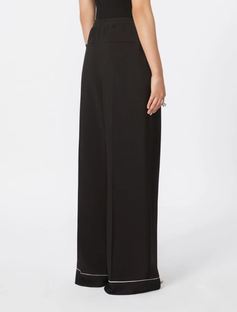 Back view of a person wearing black wide-leg trousers with white piping along the hem, elastic waistband, and a black top above.