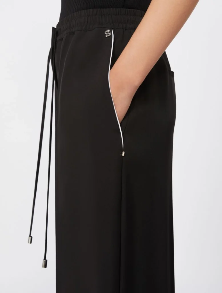 Side view of black wide-leg pants with an elastic drawstring waist, white piping along the side seam, and a hand in the pocket.