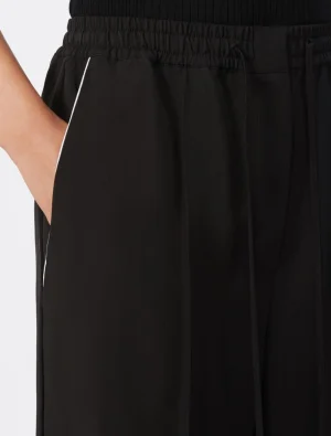 Close-up of a person’s hand resting in the pocket of black jogger pants with an elastic waistband and white piping along the side seam.] , not starting with Image