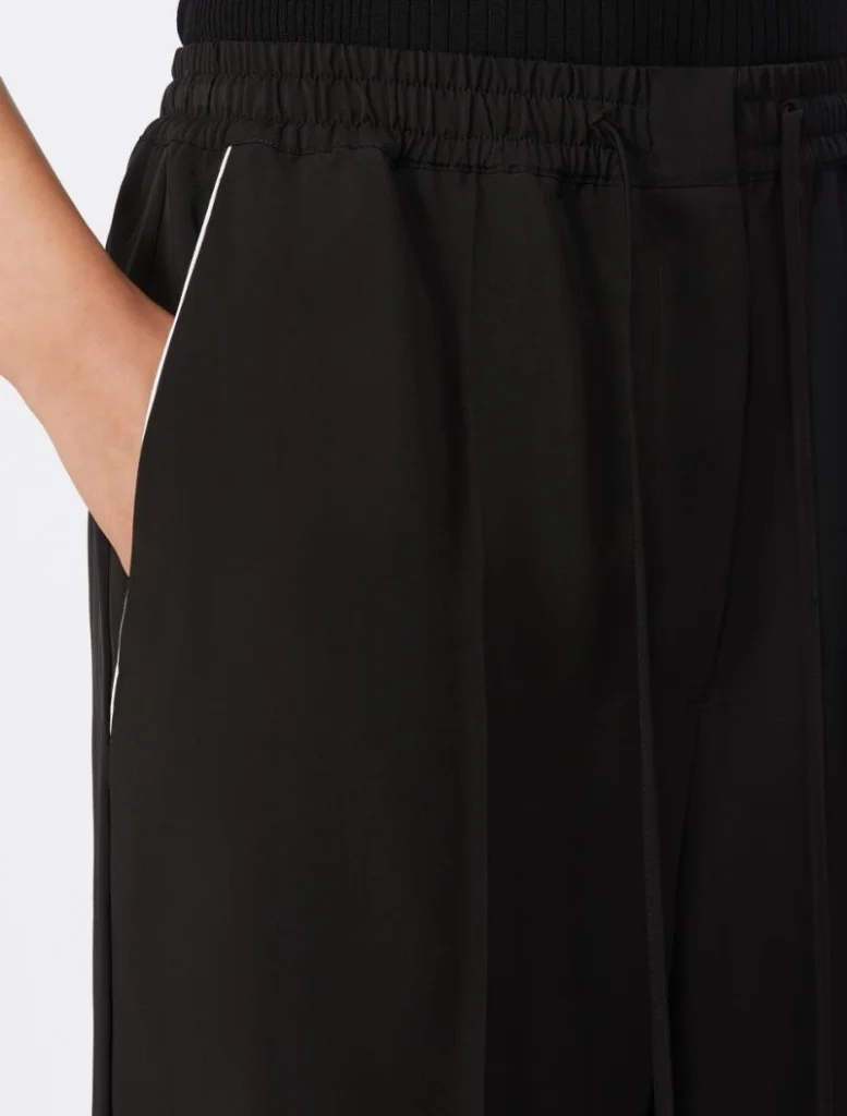 Close-up of a person’s hand resting in the pocket of black jogger pants with an elastic waistband and white piping along the side seam.] , not starting with Image