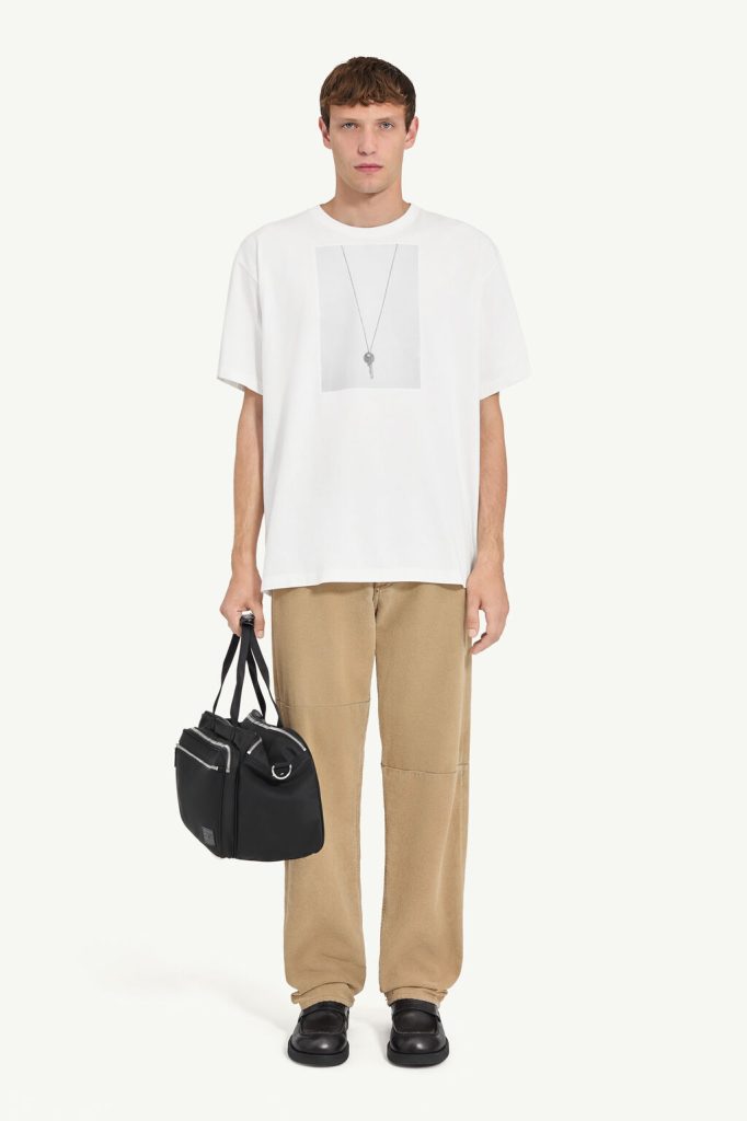 Male model wearing a white oversized T-shirt with a gray pendant graphic, tan loose-fit pants, black shoes, and holding a black handheld bag reached at side.