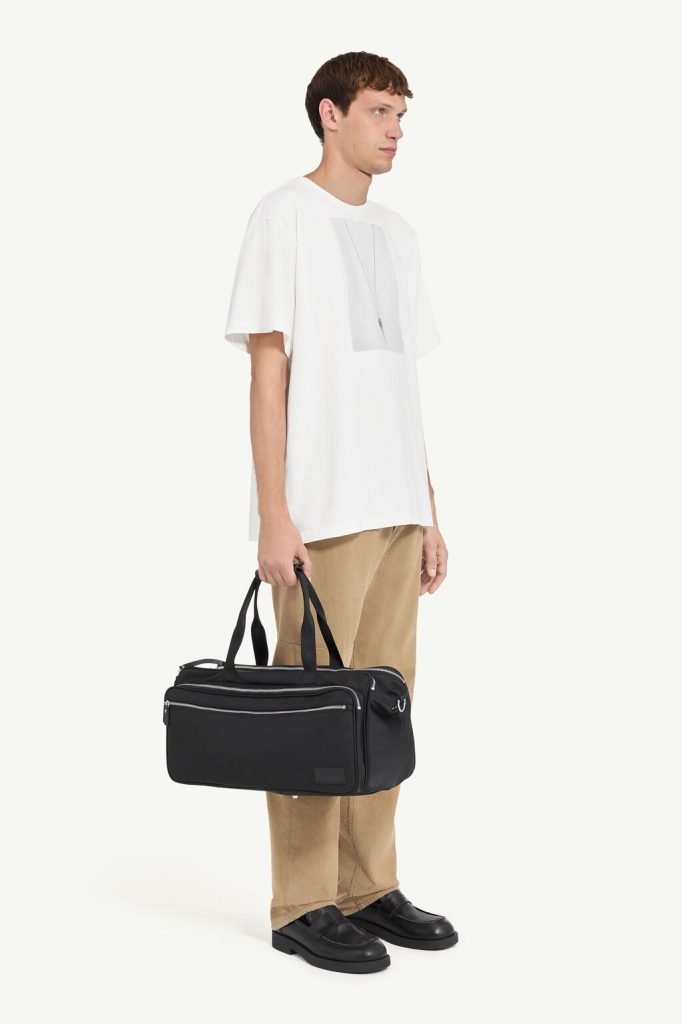 Side-view of a man in a white oversized T-shirt, tan pants, black loafers, holding a black duffel bag by the handles
