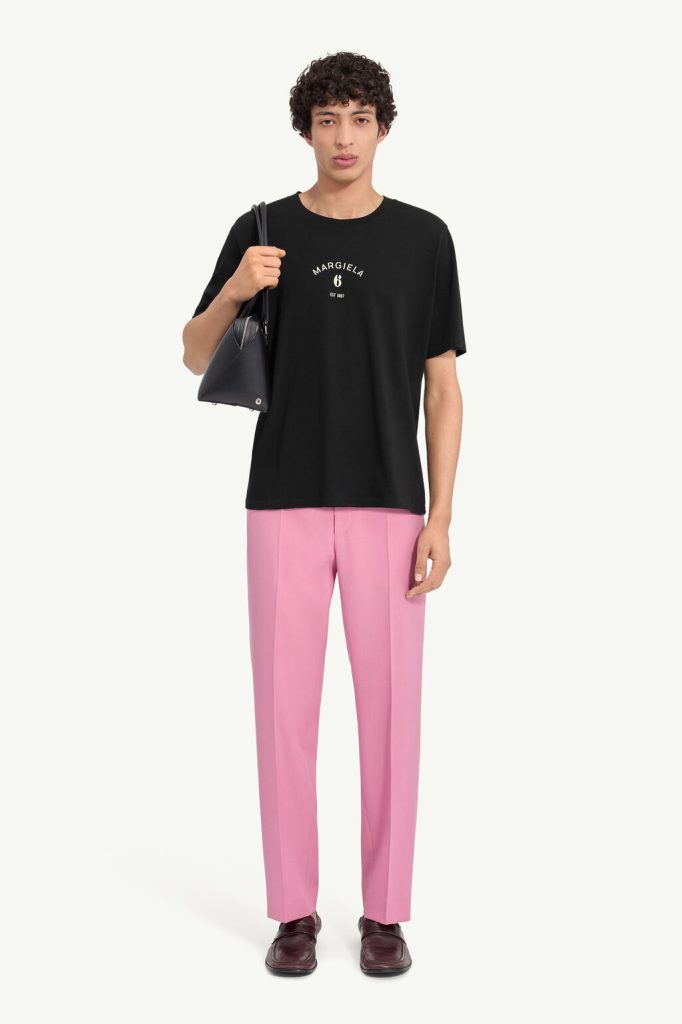 Man wearing a black T-shirt with a small white logo, pink trousers, burgundy loafers, and carrying a black handbag over his shoulder.