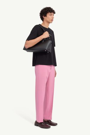Man in a black T-shirt and pink pants carries a black shoulder bag, wearing burgundy loafers (side view).