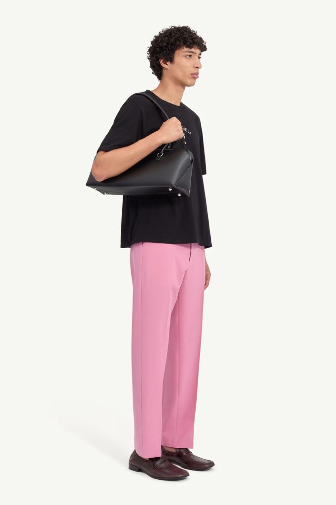 Man in a black T-shirt and pink pants carries a black shoulder bag, wearing burgundy loafers (side view).