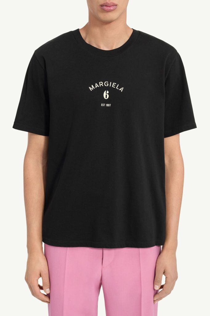 Black crew-neck T-shirt featuring a small white 'MARGIELA 6 EST 1987' logo on the chest, paired with pink trousers on a model.