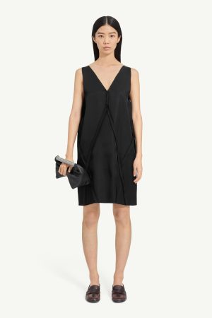 Model wearing a loose black sleeveless V-neck dress, holding a black clutch, standing against a light background.