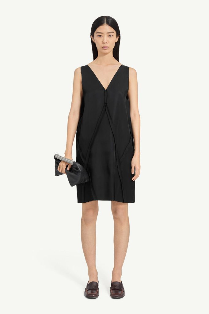 Model wearing a loose black sleeveless V-neck dress, holding a black clutch, standing against a light background.