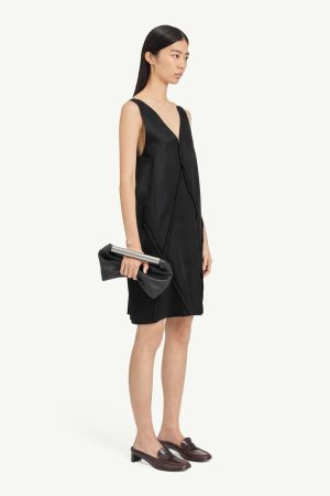 Model in a black sleeveless dress, holding a black clutch with a silver bar, wearing brown loafers, in profile view.