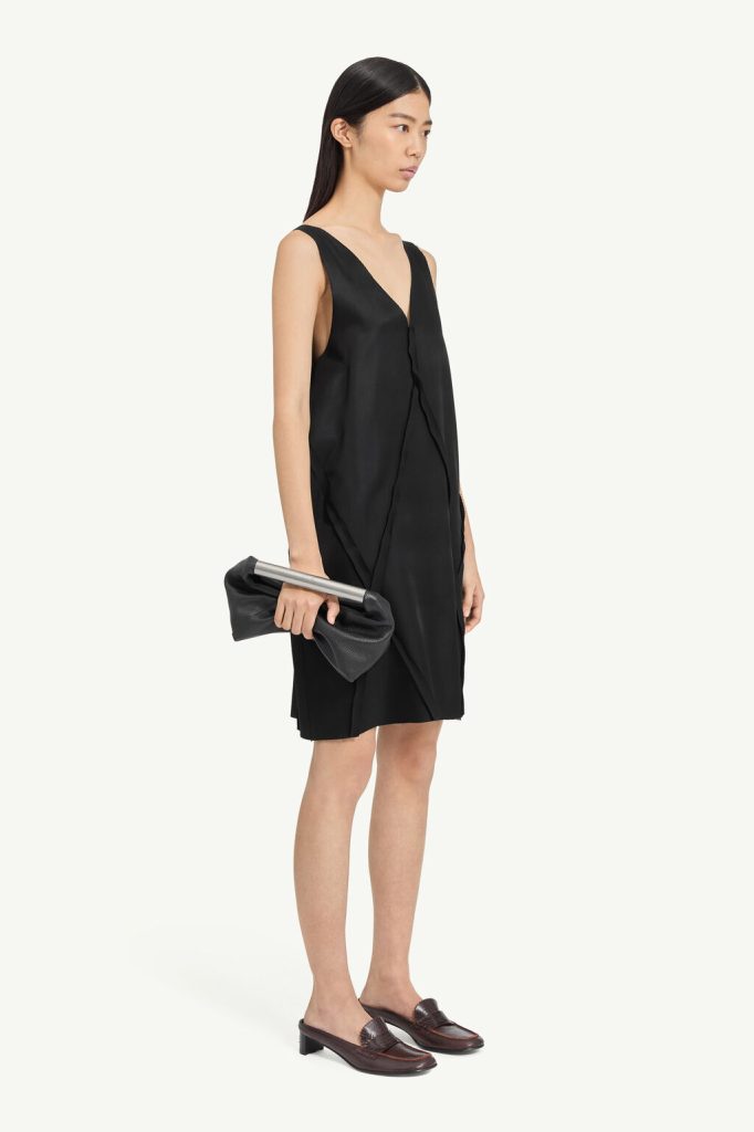 Model in a black sleeveless dress, holding a black clutch with a silver bar, wearing brown loafers, in profile view.