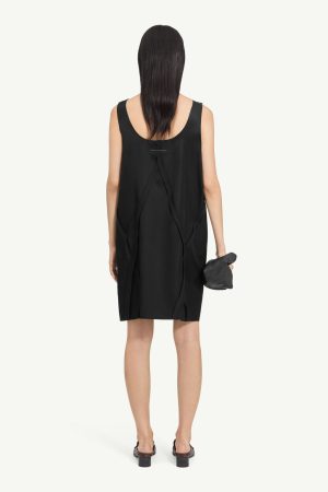 Back view of a woman in a loose black sleeveless dress with draped panels, holding a small black clutch.