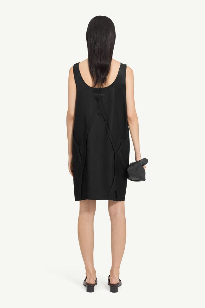 Back view of a woman in a loose black sleeveless dress with draped panels, holding a small black clutch.