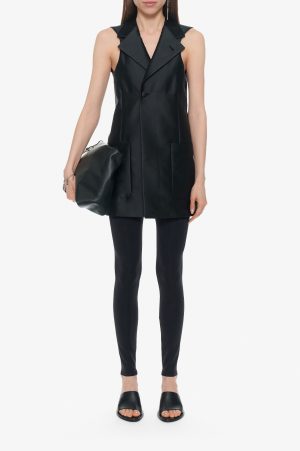 Woman modeling a black sleeveless vest with wide lapels, front pockets, paired with black leggings and slide sandals, holding a black clutch bag.