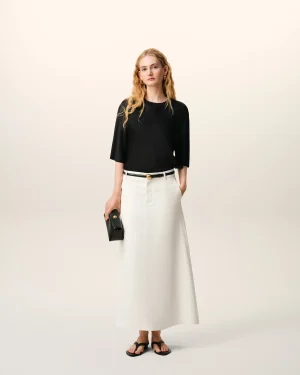 Woman standing against a light, neutral background in a black top, white midi skirt, and black sandals, holding a small black clutch.