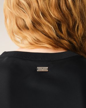 Back of a person’s neck with light blond hair, wearing a black knit top and a small rectangular metal tag with a heart symbol at center back
