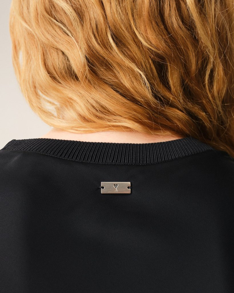 Back of a person’s neck with light blond hair, wearing a black knit top and a small rectangular metal tag with a heart symbol at center back