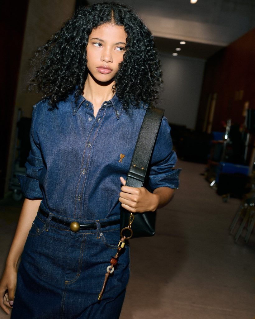 Woman with curly hair wearing a blue denim dress and a black shoulder bag, standing indoors.