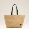 Woven straw tote bag with black handles against a light background, featuring a small heart charm on the lower right side