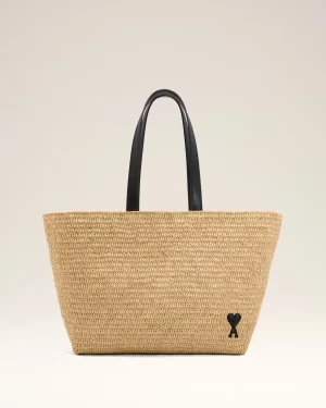 Woven straw tote bag with black handles against a light background, featuring a small heart charm on the lower right side