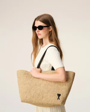 Woman in a white ribbed top wearing black sunglasses, holding a large woven straw tote bag over her shoulder.