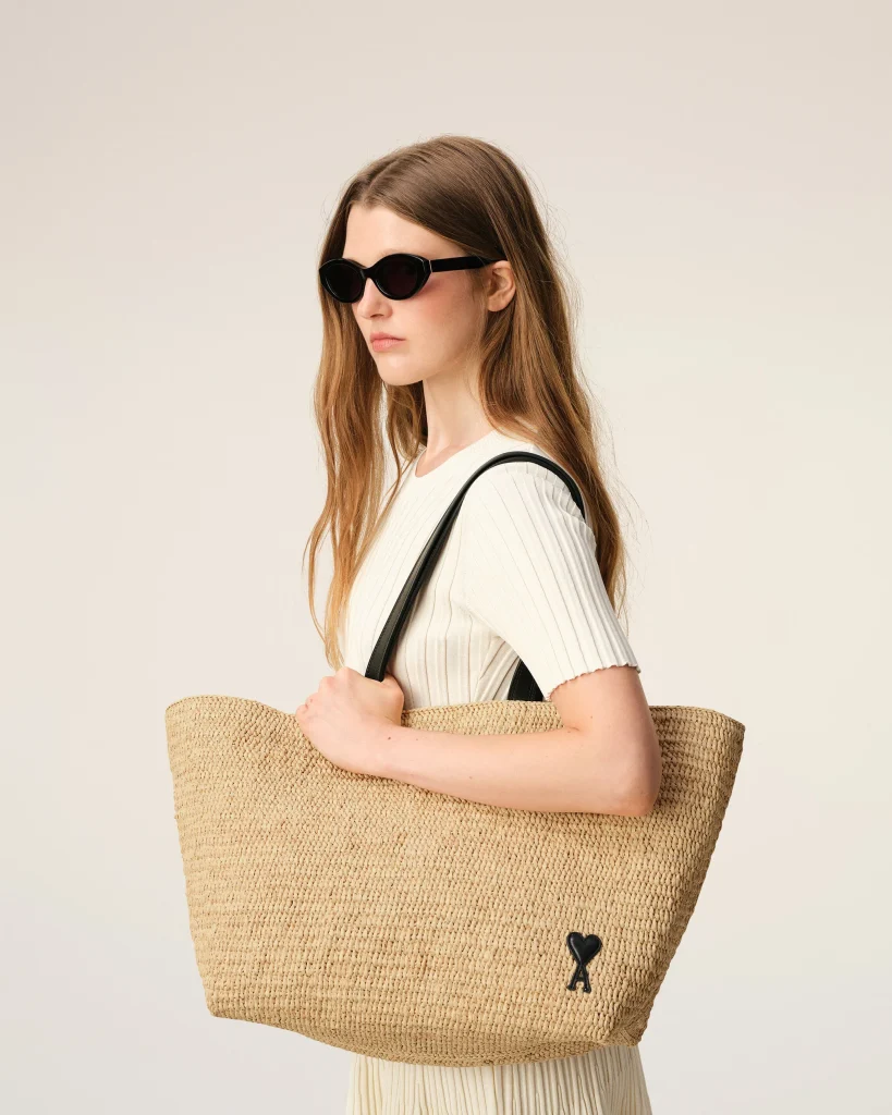 Woman in a white ribbed top wearing black sunglasses, holding a large woven straw tote bag over her shoulder.