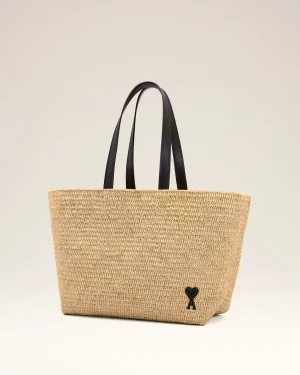 Beige woven straw tote bag with black leather handles and a small black heart charm near the bottom edge.