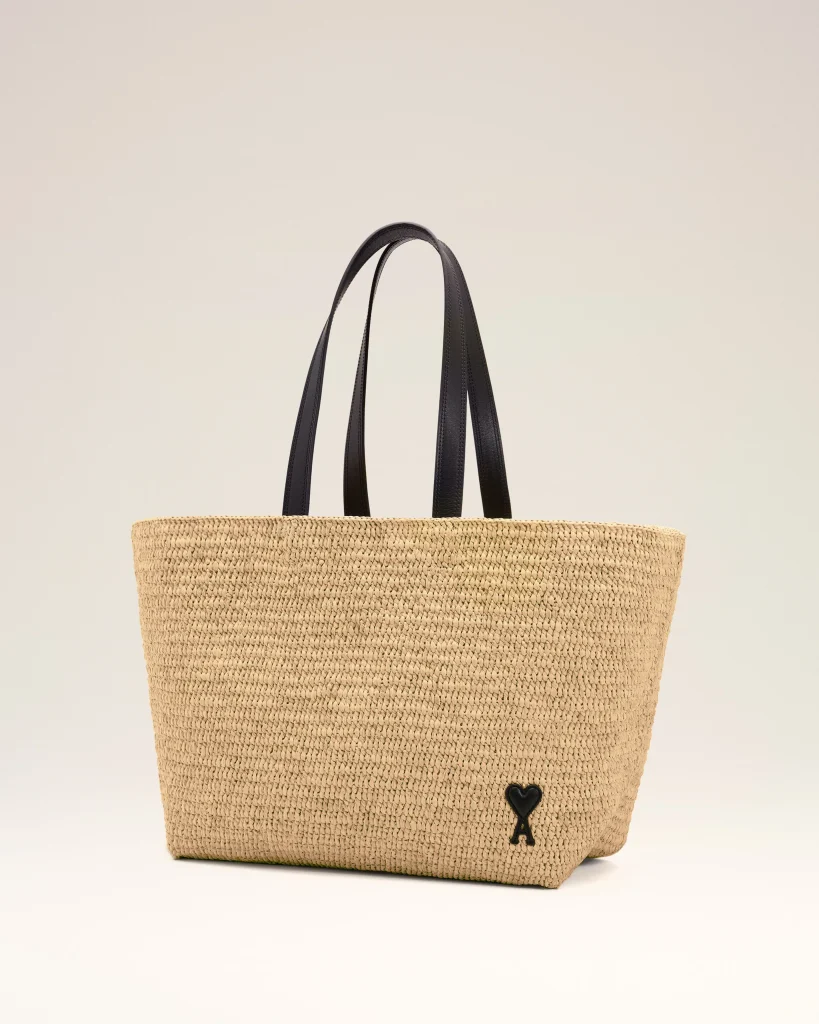 Beige woven straw tote bag with black leather handles and a small black heart charm near the bottom edge.