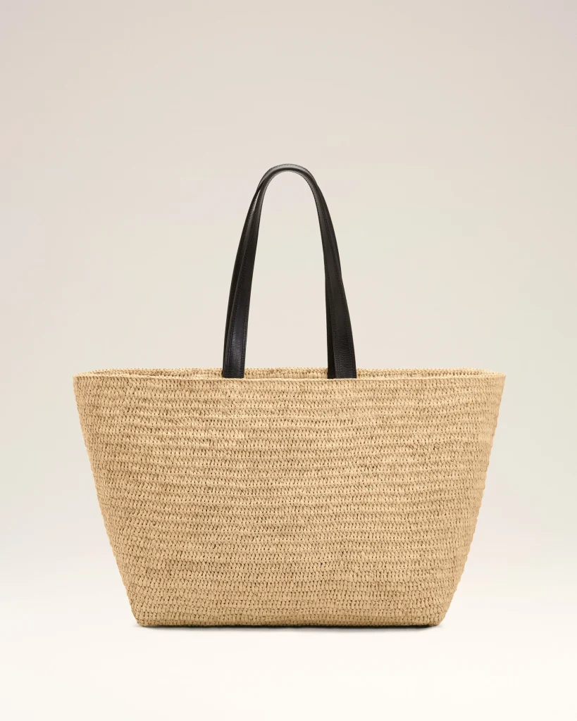 Large natural straw tote bag with two black leather handles against a light background in a minimalist style.