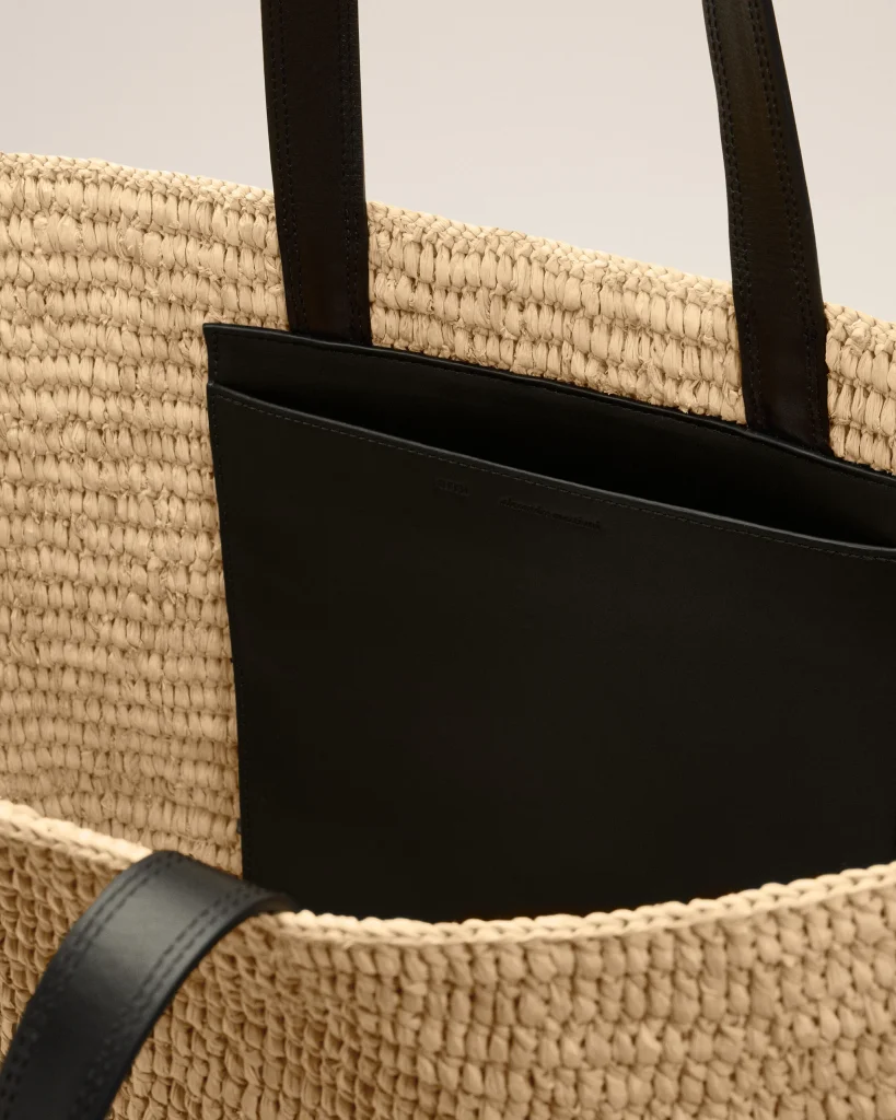 Close-up of a beige woven straw bag with black leather handles and a black front pocket edge.