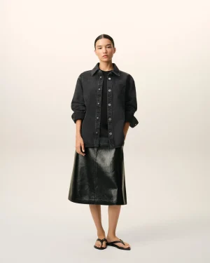 Full-length portrait of a person in a dark denim jacket over a black top and a black leather skirt, wearing black flip-flops, against a pale gradient backdrop.